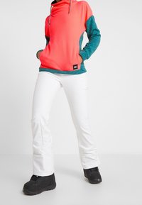 Sudadera ligera de forro polar en rosa y verde azulado con cuello de capa; combinada con pantalones blancos y botas negras. Notable contraste de color y textura.