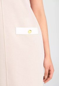 Ljusrosa klänning i tyg med en strukturerad yta, med en vit fickdetalj som är fäst med en knapp i guldton.