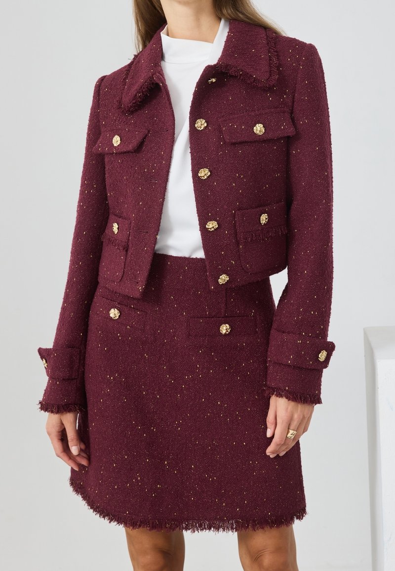 Femme portant une veste en tweed texturée bordeaux et une jupe assortie avec des boutons dorés et des bords frangés par-dessus un haut blanc.