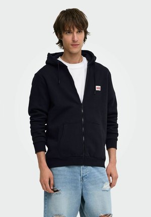 Jonge man die een zwarte hoodie met rits draagt over een wit t-shirt en lichtblauwe gescheurde jeans, staand tegen een effen lichte achtergrond.