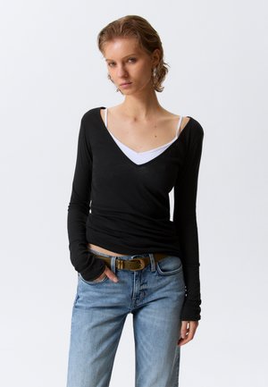 Noor lühikeste juustega, kellel on seljas must pikkade varrukatega top valge kampsunpluusiga, helesinised teksad ja pruun vöö, käed lõdvestunud.