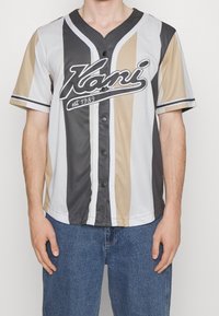 Camisa de estilo béisbol de mangas cortas en rayas gris claro, beige y gris oscuro. Presenta un escote en forma de V y un cierre de botones a presión en el centro con detalle de logotipo.