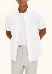 Witte short-sleeve shirt met knoopsluiting aan de voorkant, twee borstzakken en opgerolde mouwen. Draagt over een wit T-shirt en grijze broek.