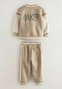 Tan värvi puuvillane treeningkostüüm valgete detailidega. Jakk on seljaosas suurte "BAKER" kirjaga. Pükstel on küljetaskud ja elastsed mansetid.