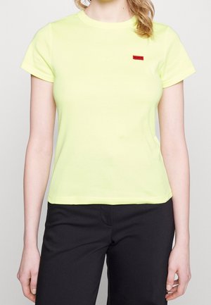 Kobieta nosząca jasnofioletowy T-shirt z krótkim rękawem z małym czerwonym logo Hugo na lewej piersi i czarne spodnie.