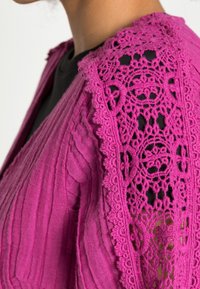 Magenta vest met een textuur van geribbelde stof en kanten details aan de mouwen, met verfijnde bloemmotieven en gekartelde randen.