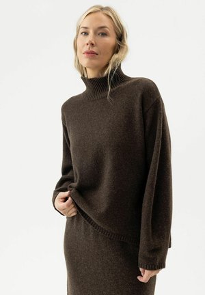 Femme blonde portant un pull à col roulé marron foncé et une jupe assortie, debout devant un fond blanc uni.
