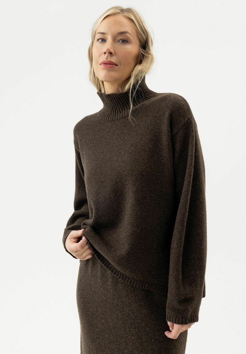 Femme blonde portant un pull à col roulé marron foncé et une jupe assortie, debout devant un fond blanc uni.