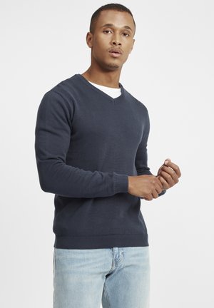 Pulover bleumarin cu decolteu în V, cu manșete și tiv plisate, fabricat din material tricotat, purtat cu blugi bleu deschis.