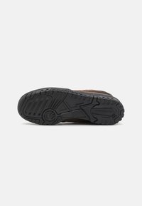 Semelle de chaussure avec un motif détaillé de bande de roulement en caoutchouc noir, comprenant des rainures circulaires et linéaires, et le logo New Balance sur un fond uni.