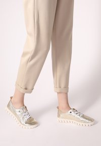 Metallisch goldene Sneakers mit weißen Schnürsenkeln und strukturierten Sohlen, kombiniert mit beigen, schmal zulaufenden Hosen. Verfügt über eine Schlaufe an der Rückseite für einfaches Anziehen.