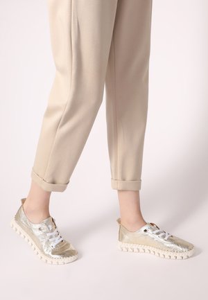 Metallisch goldene Sneakers mit weißen Schnürsenkeln und strukturierten Sohlen, kombiniert mit beigen, schmal zulaufenden Hosen. Verfügt über eine Schlaufe an der Rückseite für einfaches Anziehen.