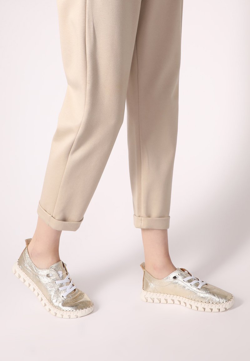 Metallisch goldene Sneakers mit weißen Schnürsenkeln und strukturierten Sohlen, kombiniert mit beigen, schmal zulaufenden Hosen. Verfügt über eine Schlaufe an der Rückseite für einfaches Anziehen.