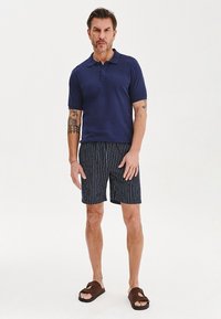 Granatowa koszulka polo z krótkim rękawem, zestawiona z ciemnymi szortami w paski. Brązowe sandały z podwójnymi paskami dopełniają strój.