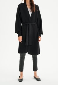 Mustat ribatut kardiganit, joissa on vyötäröllä vyö, leveät hihat ja avonainen etuosa. Yhdistetty istuviin mustiin nahkahousuihin ja tasapohjaisiin kenkiin.