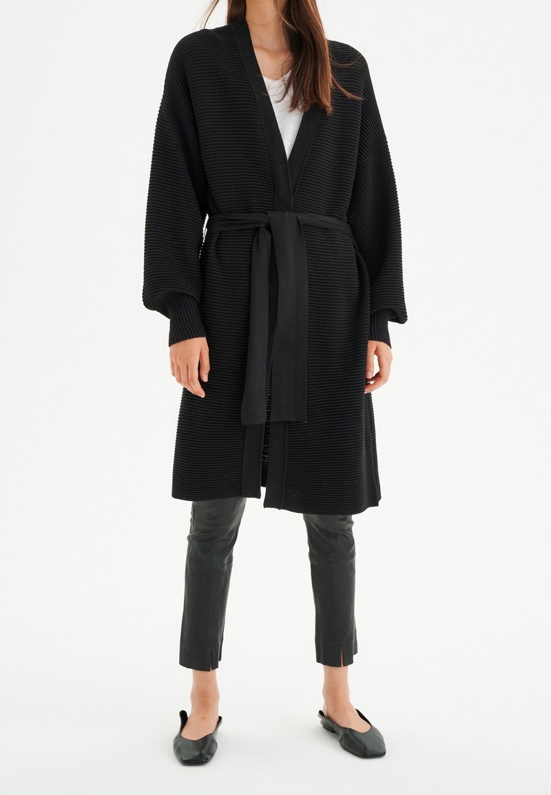 Mustat ribatut kardiganit, joissa on vyötäröllä vyö, leveät hihat ja avonainen etuosa. Yhdistetty istuviin mustiin nahkahousuihin ja tasapohjaisiin kenkiin.