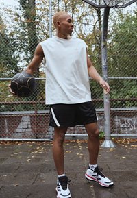 Top atletico bianco senza maniche, pantaloni corti neri con accenti bianchi, scarpe da ginnastica nere e rosse. Tiene in mano un pallone da basket nero. Ambientazione di un campo all'aperto.