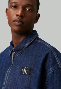 Denim jakna v temno modri barvi z zadrgo, koničastim ovratnikom in našitkom blagovne znamke Calvin Klein Jeans. Ima vidne šive.