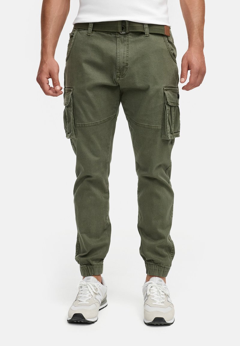 Homme portant un pantalon cargo vert olive avec des poignets élastiques et une ceinture verte, assorti à des baskets blanches.