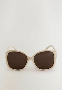 Beige oversized solbriller med mørkebrune linser, en glat plastisk ramme og subtile hardware-detaljer på siderne.