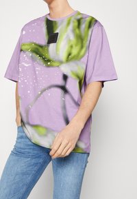 Lila bomullströja med ett stort, abstrakt grönt och vitt tryck, korta ärmar och en liten svart etikett nära halslinningen.