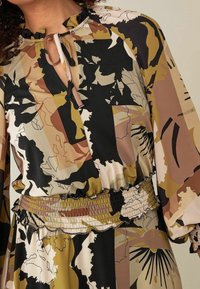 Robe imprimée en marron, noir et crème. Présente un décolleté en trou de clé, un col à volants et une taille élastiquée avec un détail texturé.