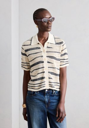 Žena nosiaca pruhovanú krátkym rukávom pletenú košeľu na gombíky, modré džínsy, slnečné okuliare, zlaté kruhové náušnice a široký zlatý náramok, stojaca v interiéri.