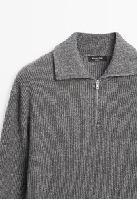 Pull en maille côtelée gris avec un col zippé, présentant un design en tricot texturé. Étiquette de la marque visible à l'intérieur du col.