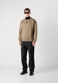 Bež pulover s majhnim logotipom, črna turtleneck majica spodaj, črne kavbojke in črne čevlje; model nosi temne sončne očala.