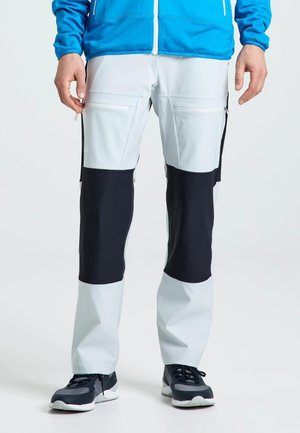 Modelo con pantalones cargo gris claro con paneles negros en las rodillas, una chaqueta azul y zapatillas oscuras sobre un fondo liso.