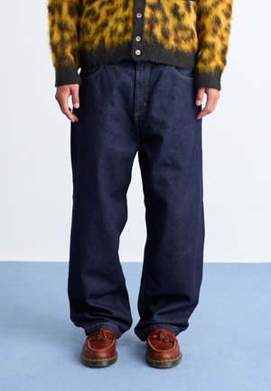 Blue denim wide-leg pants with front pockets and yellow stitching, paired with brown moccasin-style shoes. The background is a light color.