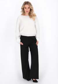 Weißer, locker sitzender Pullover mit langen Ärmeln und geripptem Saum, kombiniert mit hoch taillierten, weit geschnittenen schwarzen Hosen. Schwarze Pumps runden das Outfit ab.