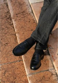 Black leather loafers with a smooth finish, side stitching, and a raised seam detail, worn with black ribbed socks and dark jeans.