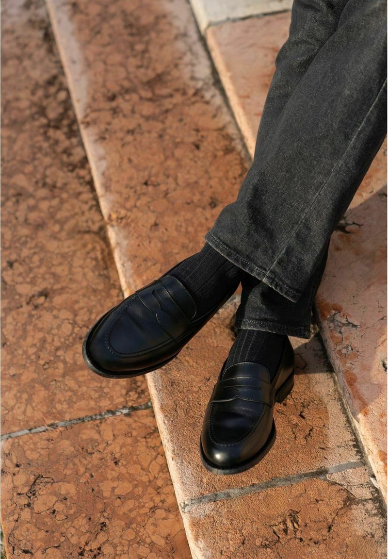 Black leather loafers with a smooth finish, side stitching, and a raised seam detail, worn with black ribbed socks and dark jeans.