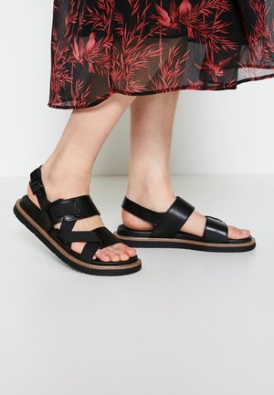 Jambes portant des sandales ouvertes en cuir et tissu noir avec boucles, debout sur un sol clair, ourlet d'une jupe noire transparente avec motif floral rouge visible.