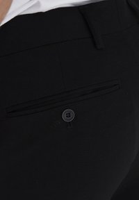 Schwarze Hose mit glattem Stoff, ausgestattet mit einer einzelnen Gesäßtasche und einem schwarzen Knopf mit der Aufschrift "FRIDAY CASUAL".