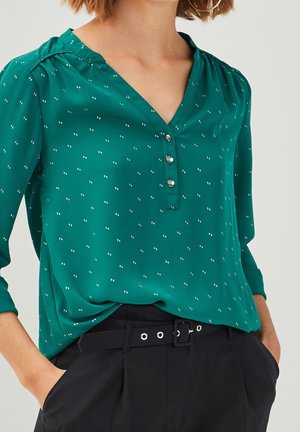 Femme portant un chemisier vert à manches longues avec de petits pois blancs et trois boutons, associé à un pantalon noir ceinturé et mains dans les poches.