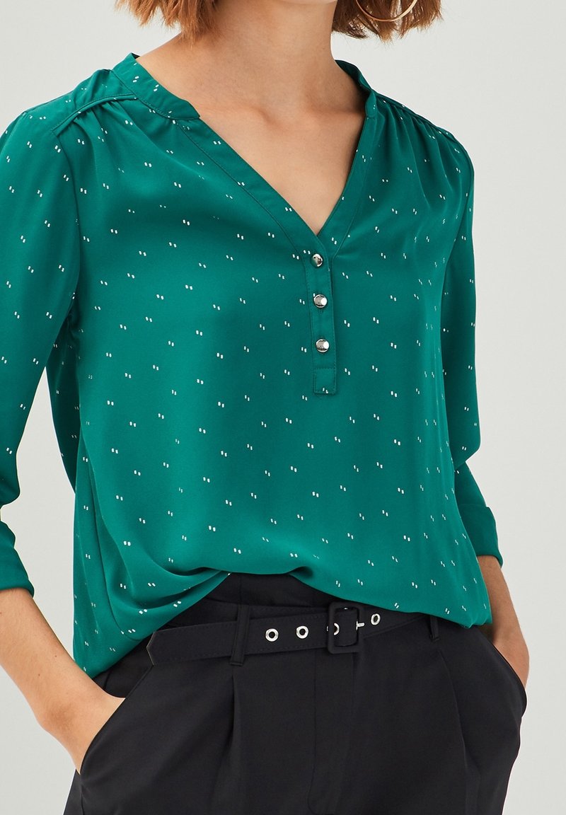 Femme portant un chemisier vert à manches longues avec de petits pois blancs et trois boutons, associé à un pantalon noir ceinturé et mains dans les poches.
