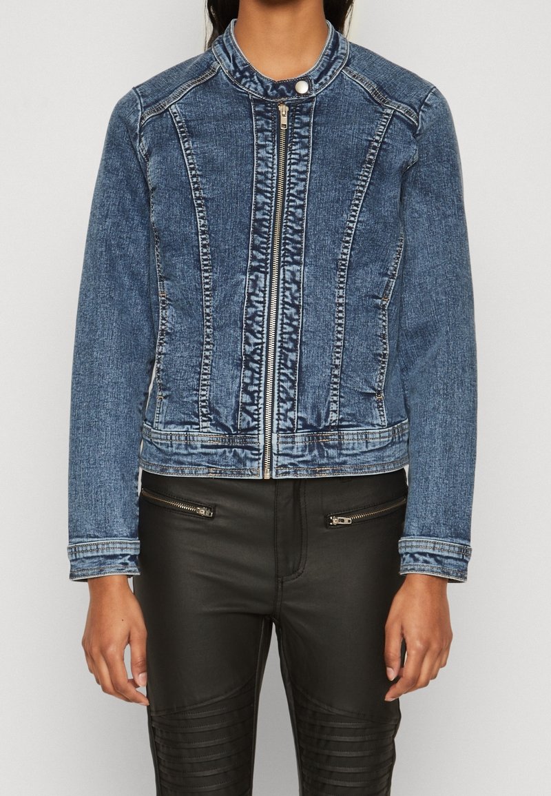 Blå denimjacka med dragkedja fram, rund krage och framträdande sömdetaljer. Buren över svarta strukturerade byxor med dragkedjefickor.