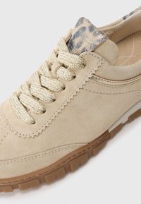 Beige semsket sko med teksturert overflate, kontrasterende lys brun gummisåle, kremfargede snørebånd, og en mønstrete tunge med leopardmønster.