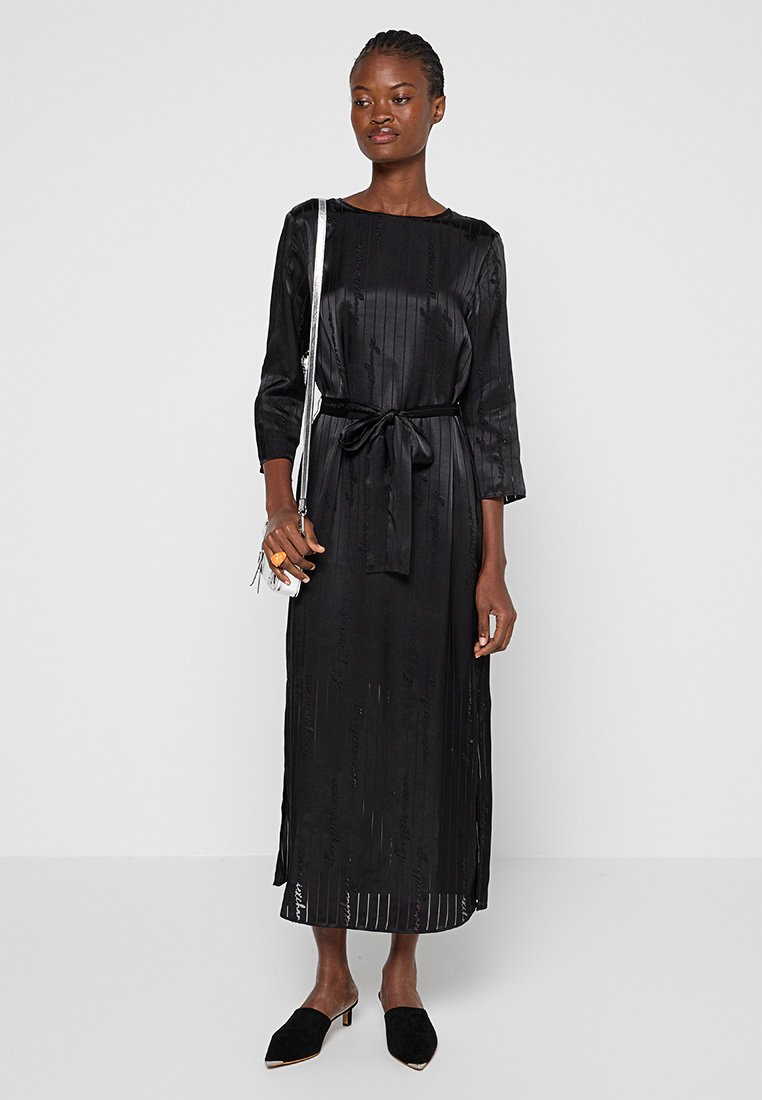 Černé saténové šaty s dlouhým rukávem, vertikálním žebrovaným vzorem, opaskem v pase, bočními rozparky a uvolněným střihem, v kombinaci s černými muly a stříbrnou kabelkou.