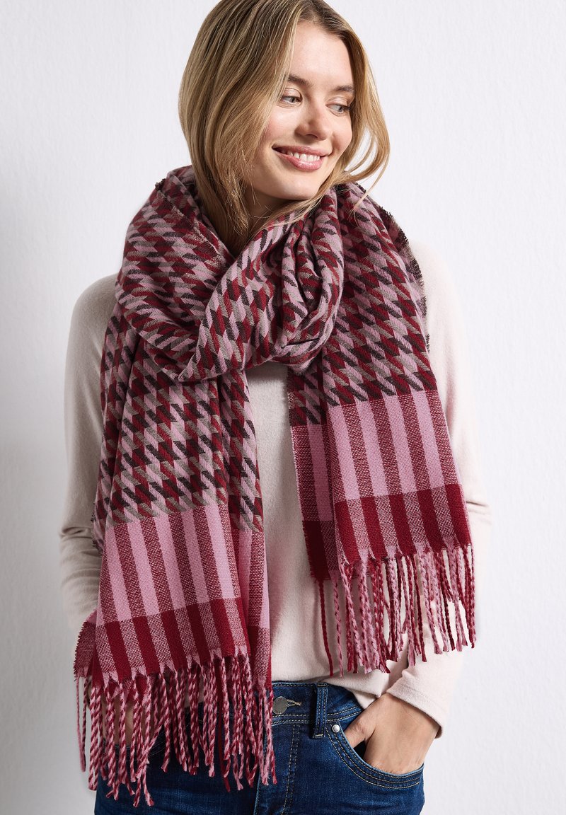 Jeune femme portant un pull clair et un jean bleu, avec une grande écharpe rouge et rose à motifs enroulée autour du cou et des épaules.