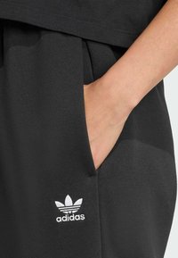Crne sportske hlače s teksturiranim materijalom, s bočnom džepom i bijelim Adidas logotipom na donjem dijelu nogavice.