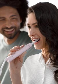 Spazzolino elettrico in plastica rosa chiaro, design snodato, con testina argentata e una luce verde indicativa, tenuto da una donna.