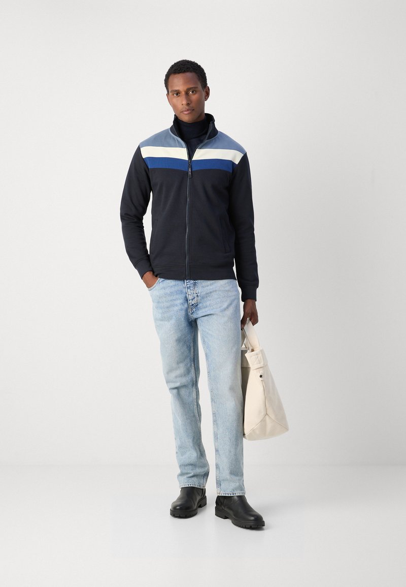 Veste zippée navy avec des rayures horizontales bleues, blanches et grises, associée à un jean bleu clair et des chaussures noires. Tenant un sac de couleur claire.