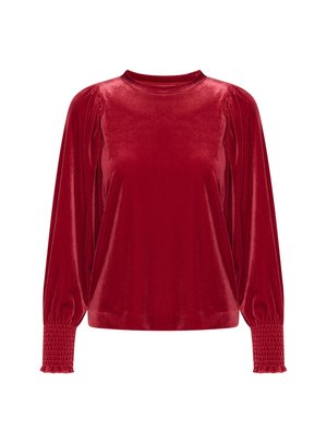Blusa lunga in velluto rosso con colletto alto, spalle arricciate e polsini smock. La texture liscia esalta il colore ricco.