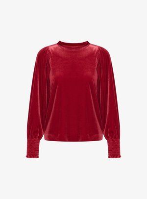 Blusa lunga in velluto rosso con colletto alto, spalle arricciate e polsini smock. La texture liscia esalta il colore ricco.