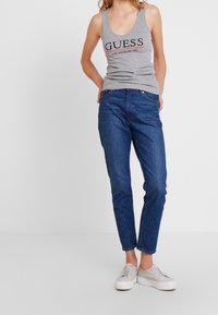 Canottiera grigia con logo "GUESS", abbinata a jeans in denim blu. Le scarpe sono sneakers grigie chiare. Design semplice, vestibilità casual, senza fantasie.