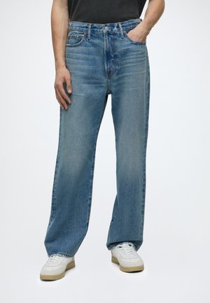 Homme portant un jean bleu ample et des baskets blanches, debout avec une main dans la poche et l'autre main détendue le long du corps.