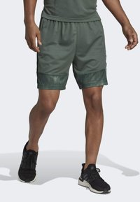 Man i mörkgröna träningsshorts med svarta detaljer och svarta löparskor, stående med ett ben framåt mot en vit bakgrund.